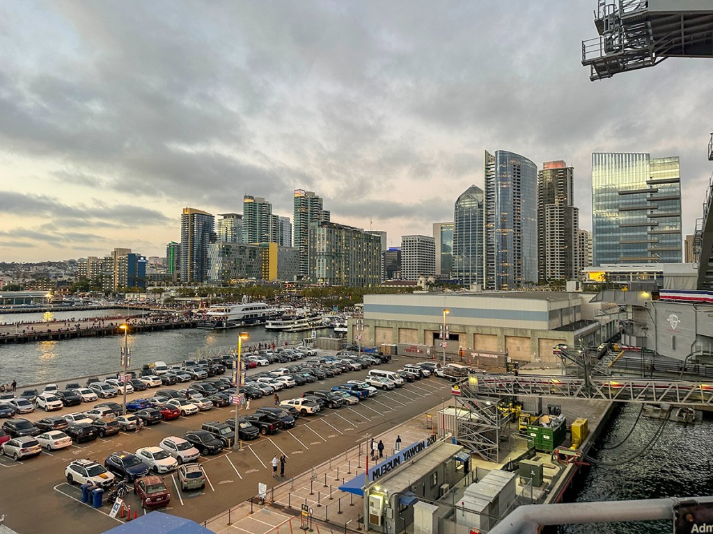 A stunning evening view of San Diego's downtown waterfront, featuring modern high-rise buildings and a bustling harbor filled with boats and activity. The iconic skyline showcases sleek, glass-covered skyscrapers, while the USS Midway Museum and surrounding piers add a touch of history and maritime culture. Cars fill the waterfront parking lot, reflecting the city's vibrant energy. Soft clouds and the fading daylight create a serene backdrop for this lively urban scene. Perfect for themes related to travel, city life, and the beauty of San Diego.