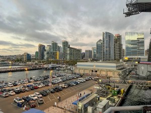 A stunning evening view of San Diego's downtown waterfront, featuring modern high-rise buildings and a bustling harbor filled with boats and activity. The iconic skyline showcases sleek, glass-covered skyscrapers, while the USS Midway Museum and surrounding piers add a touch of history and maritime culture. Cars fill the waterfront parking lot, reflecting the city's vibrant energy. Soft clouds and the fading daylight create a serene backdrop for this lively urban scene. Perfect for themes related to travel, city life, and the beauty of San Diego.