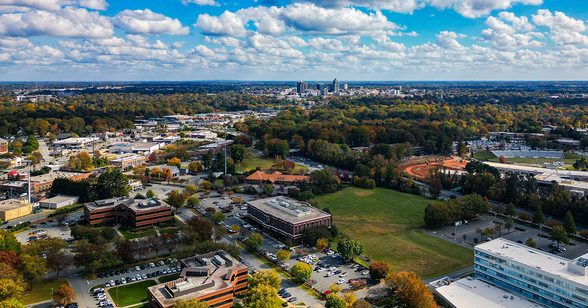 Aerial view of Raleigh, North Carolina, showcasing urban landscape and lush greenery under a vibrant blue sky with fluffy clouds.