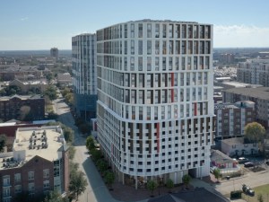 Aerial rendering of The Penny on College Main, a 20-story building with white, glass and red and black exterior. The high-rise is surrounded by other buildings.