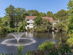 Apartment building with brown roof behind a pond with water feature and surrounded by trees