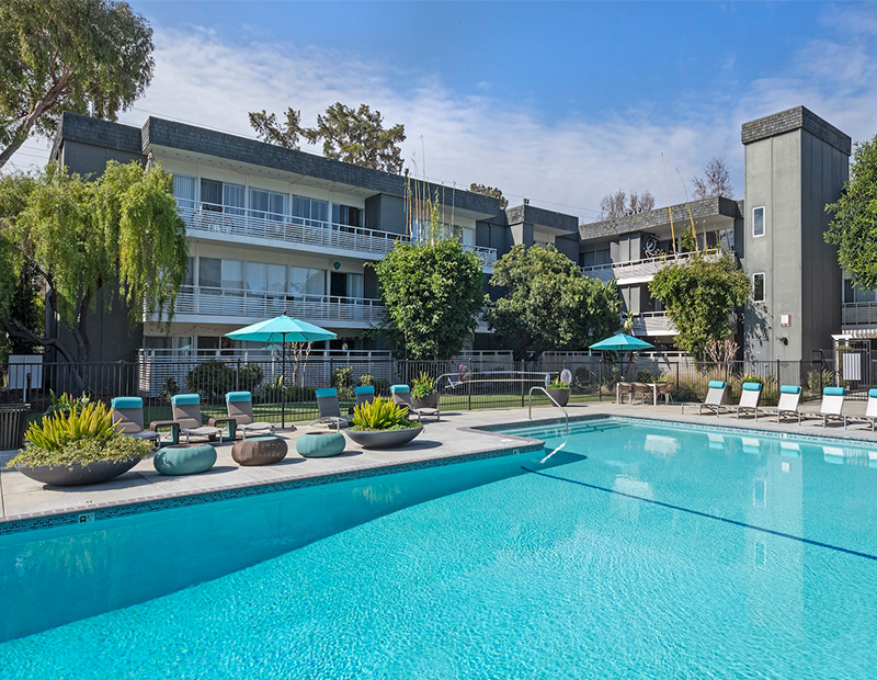 Photo of low-rise apartment building with pool, deck chairs and beach umbrellas in the foreground.