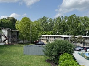 Two low-rise , off-white apartment buildings beneath trees with bushes and a lawn in the foreground