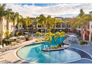 S-shaped swimming pool surrounded by deck chairs and palm trees in front a two-level light beige apartment bui