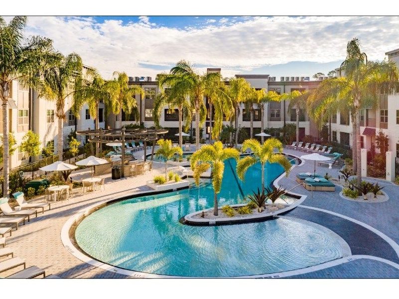 S-shaped swimming pool surrounded by deck chairs and palm trees in front a two-level light beige apartment bui