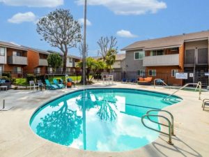 Two low-level apartment buildings with sloped roofs surrounding a small kidney-shaped pools in Southern California
