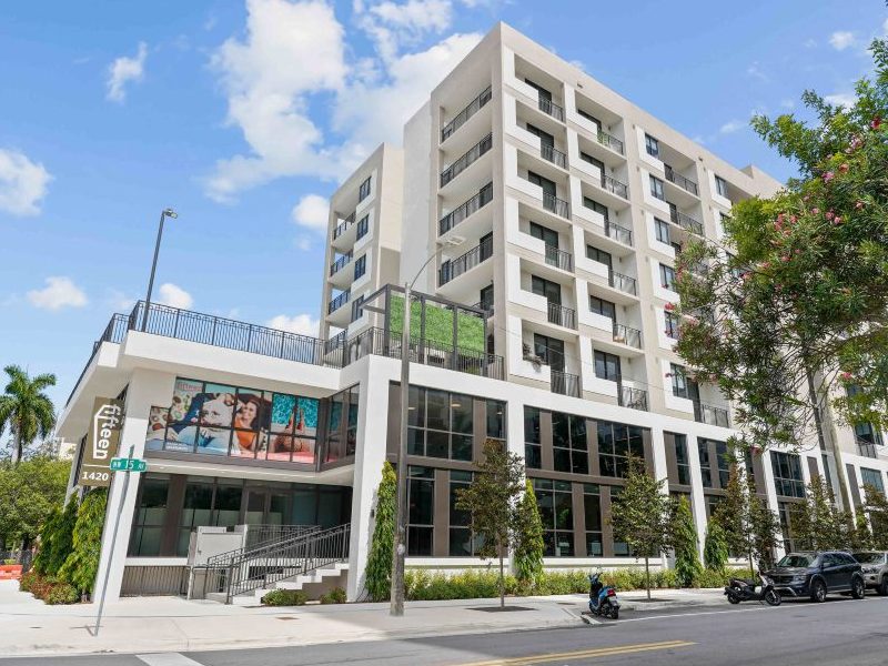 Street-level view of off-white apartment building with six levels of apartments on a two-story platform. Tree visible in right foreground, palm tree in left background