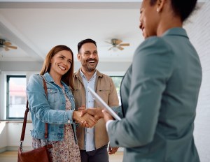 A smiling couple shakes hands with a realtor, both expressing happiness. The setting is a bright, modern space with ceiling fans and large windows.