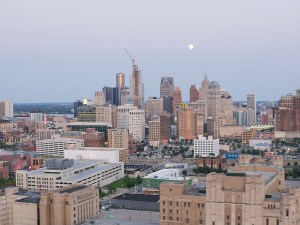 The Detroit, Michigan skyline showcases towering buildings, construction cranes, and a full moon glowing in a serene, early evening sky. The view highlights the city's dynamic urban landscape and architectural diversity.