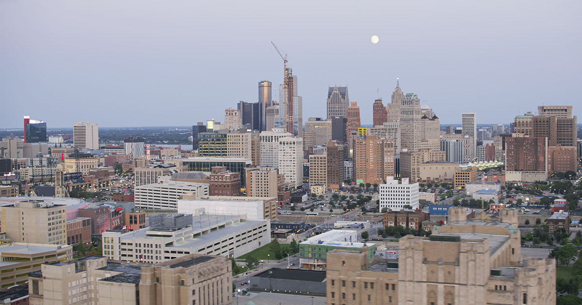 The Detroit, Michigan skyline showcases towering buildings, construction cranes, and a full moon glowing in a serene, early evening sky. The view highlights the city's dynamic urban landscape and architectural diversity.