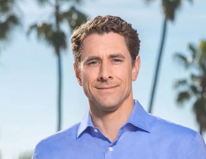 Smiling brown-haired man wearing an open-necked blue shirt with palm trees in the background