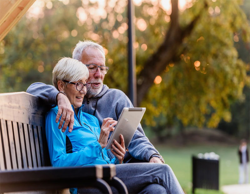 Smiling senior active couple sitting on the bench looking at tablet computer.