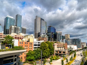View of downtown Seattle, Washington, United States, captured from Overlook Walk. The elevated pedestrian pathway offers a sweeping panorama of the city's skyline, including modern skyscrapers and waterfront buildings, framed by greenery and a partly cloudy summer sky