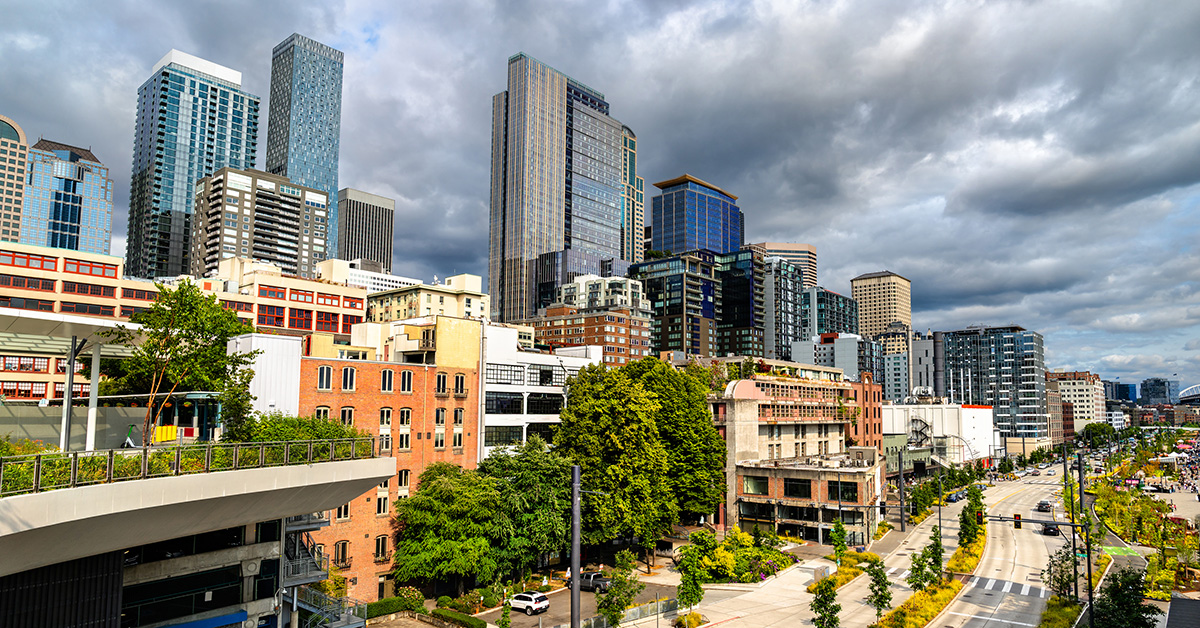 View of downtown Seattle, Washington, United States, captured from Overlook Walk. The elevated pedestrian pathway offers a sweeping panorama of the city's skyline, including modern skyscrapers and waterfront buildings, framed by greenery and a partly cloudy summer sky