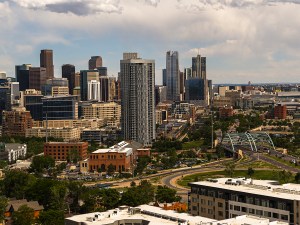 Aerial view of downtown Denver, Colorado showing the city skyline with skyscrapers, office buildings, and residential towers. The Millennium Bridge is visible connecting neighborhoods with the business district. The image captures both the dense urban core with high-rise buildings and surrounding residential areas with parks and green spaces. Partly cloudy sky creates natural lighting across the cityscape. Wide shot perspective showcases the contrast between commercial downtown and neighboring residential districts.
