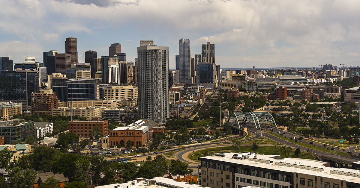 Aerial view of downtown Denver, Colorado showing the city skyline with skyscrapers, office buildings, and residential towers. The Millennium Bridge is visible connecting neighborhoods with the business district. The image captures both the dense urban core with high-rise buildings and surrounding residential areas with parks and green spaces. Partly cloudy sky creates natural lighting across the cityscape. Wide shot perspective showcases the contrast between commercial downtown and neighboring residential districts.