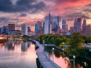 Philadelphia, Pennsylvania, USA, at sunrise. The skyline features modern skyscrapers reflecting on the Schuylkill River. A winding paved path for walking and cycling lines the river's edge. The sky is adorned with colorful clouds, creating a serene atmosphere. Lush green trees border the river, providing contrast to the urban landscape. The scene captures the beautiful blend of natural and architectural elements at the break of day.