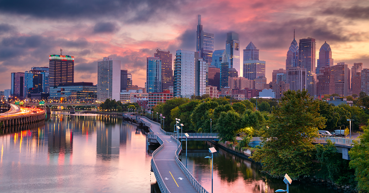 Philadelphia, Pennsylvania, USA, at sunrise. The skyline features modern skyscrapers reflecting on the Schuylkill River. A winding paved path for walking and cycling lines the river's edge. The sky is adorned with colorful clouds, creating a serene atmosphere. Lush green trees border the river, providing contrast to the urban landscape. The scene captures the beautiful blend of natural and architectural elements at the break of day.