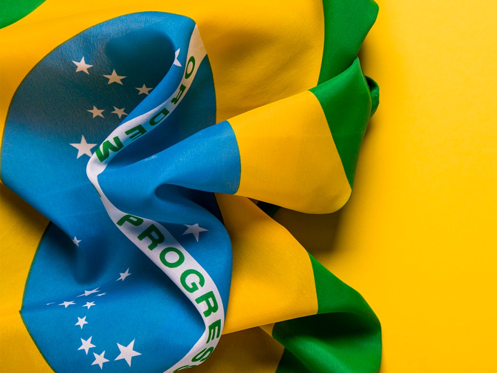 A photograph of the Brazilian national flag laying on a plain yellow background. There is space for text.