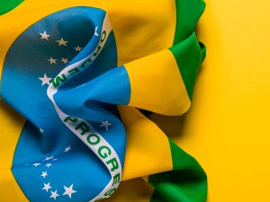 A photograph of the Brazilian national flag laying on a plain yellow background. There is space for text.