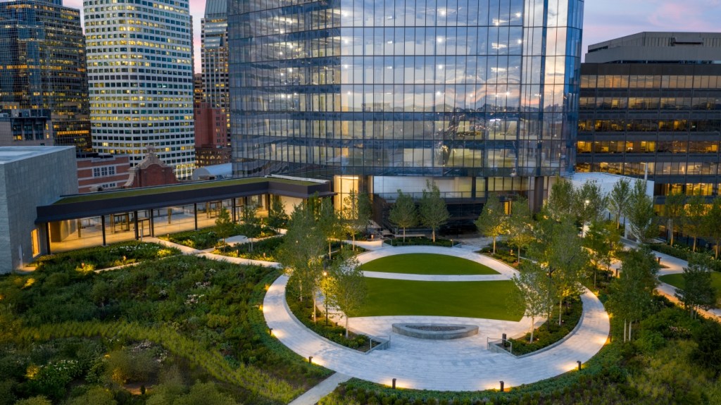 Aerial view of a modern urban rooftop garden at dusk, featuring a circular grassy area, surrounded by a walkway and trees, with city skyscrapers nearby.