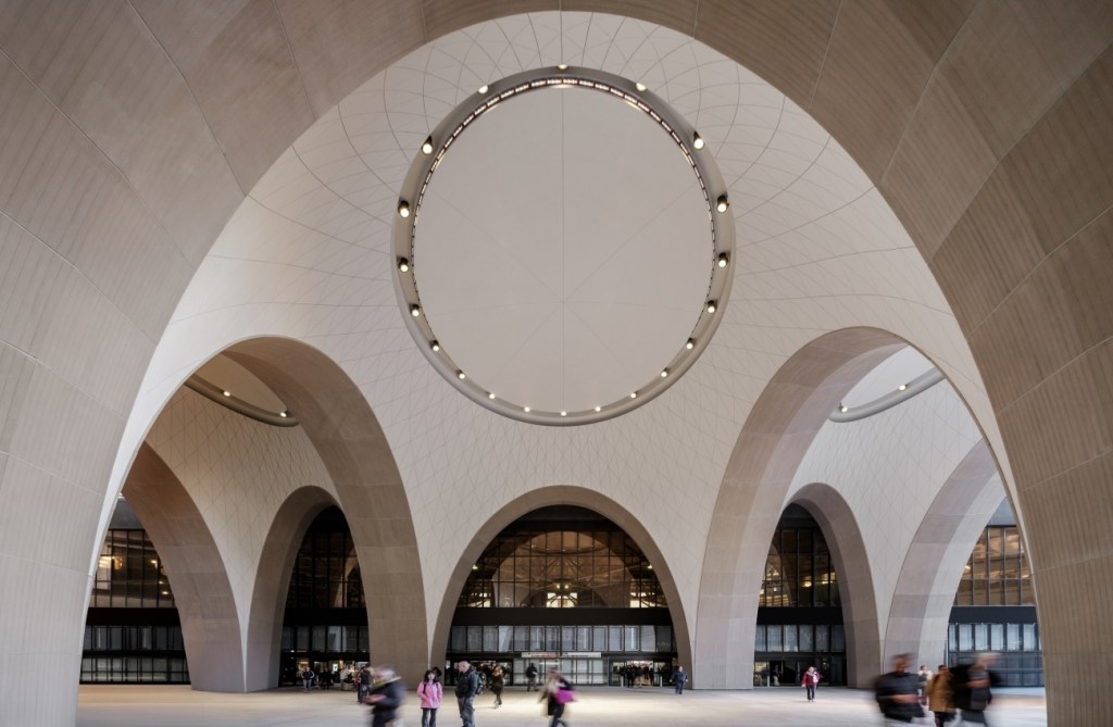Grand interior space with towering, symmetrical arches and a circular pattern overhead. People walking below add a sense of scale and liveliness.