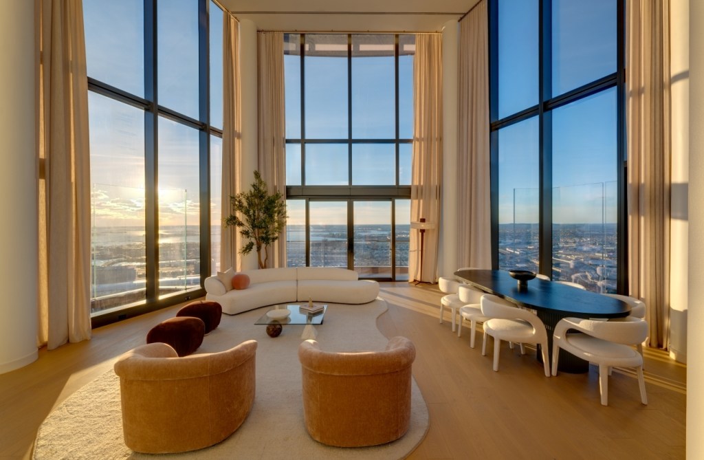 Luxurious living room with floor-to-ceiling windows showcasing a sunset view. Features include a curved sofa, armchairs, and a dining table with chairs.