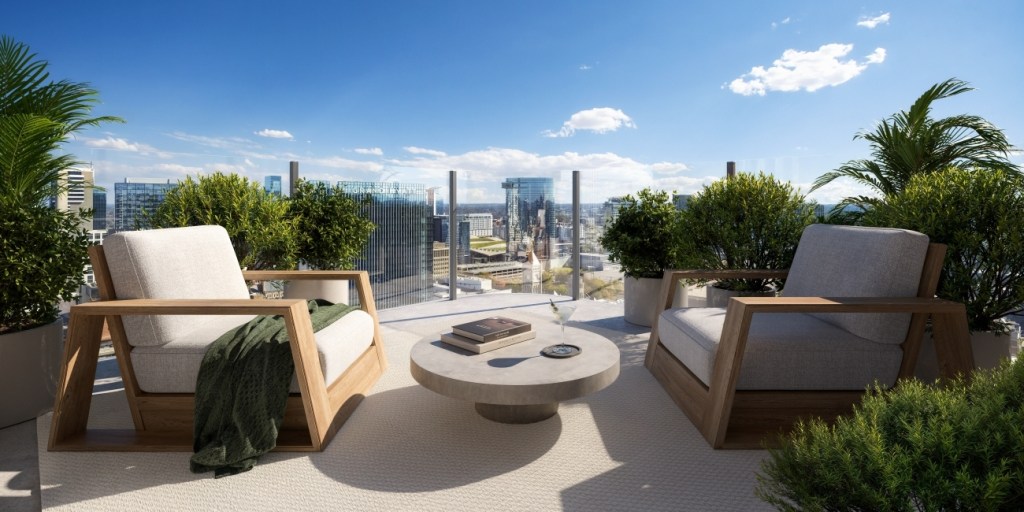 Rooftop terrace with two wooden armchairs surrounding a round table, lush plants, and a panoramic city view under a clear blue sky. Peaceful ambiance.