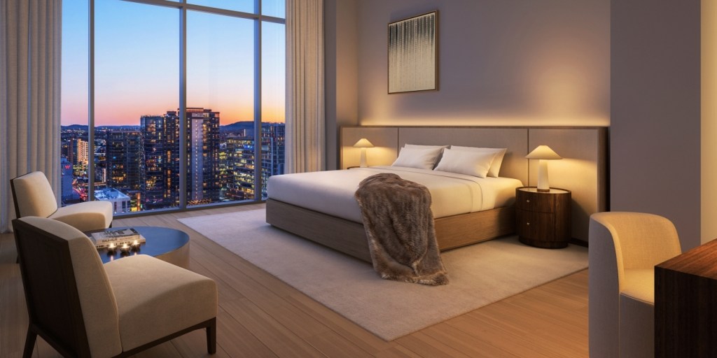 Elegant hotel room with a large bed, plush fur throw, and soft lighting. Floor-to-ceiling window reveals a cityscape at dusk, creating a warm, inviting ambiance.
