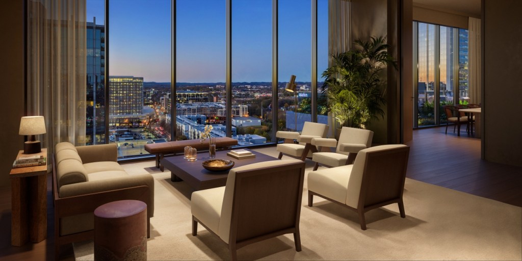 Sophisticated living room with modern furniture and large windows offering a cityscape view at dusk. Warm lighting creates a cozy, inviting atmosphere.