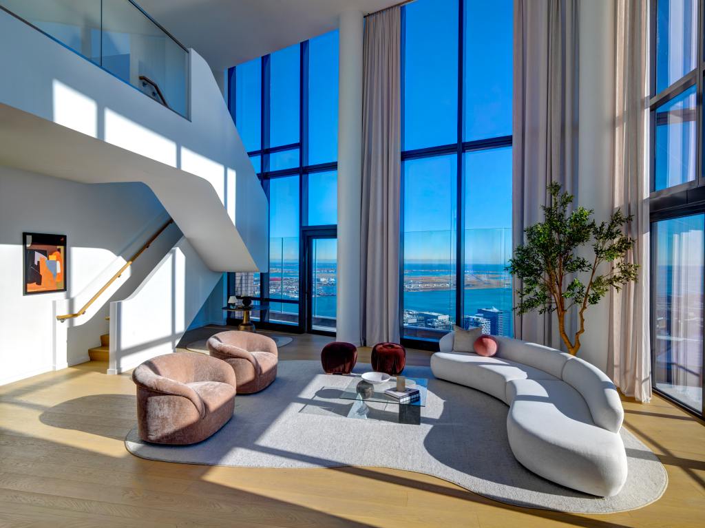 Spacious modern living room with floor-to-ceiling windows offering a coastal view. Features plush chairs, a curved sofa, a glass table, and a staircase.