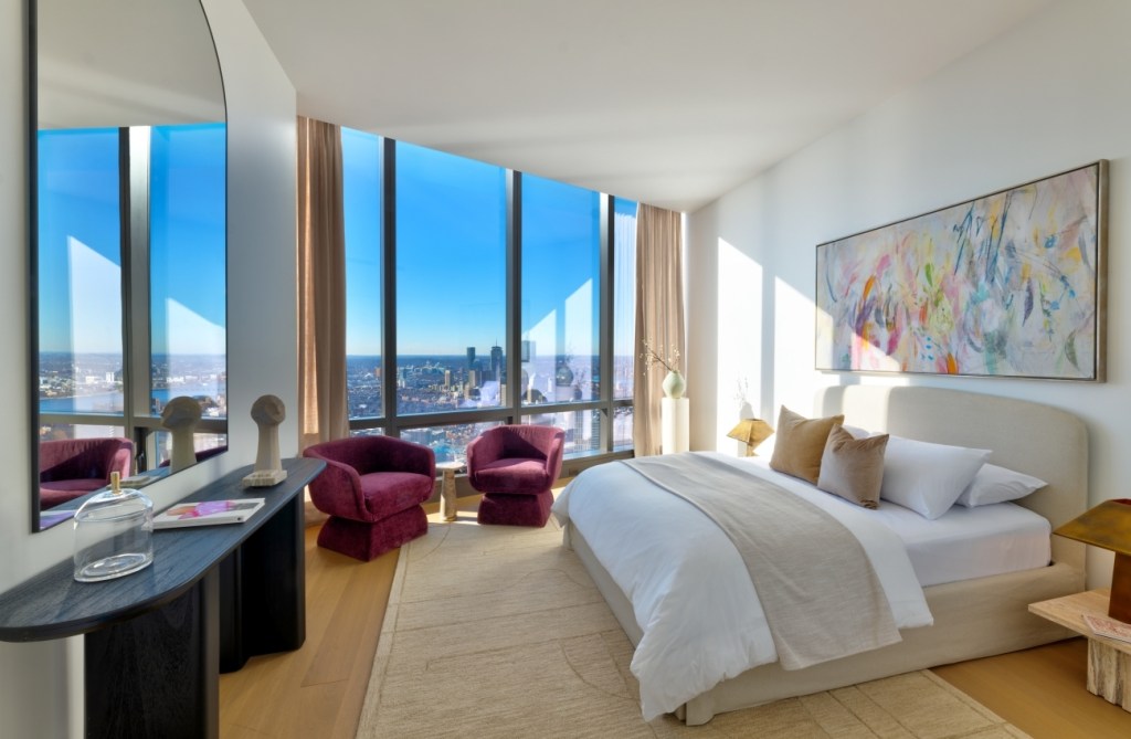 Modern bedroom with a large bed, abstract art, and burgundy chairs by floor-to-ceiling windows showing a city skyline. Bright and serene ambiance.