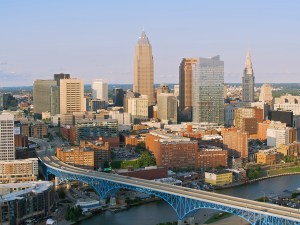 An aerial view of Cleveland, Ohio, featuring a bustling downtown skyline, iconic blue bridge, and clear skies. The urban landscape highlights the city's architecture and riverside setting on a sunny day.