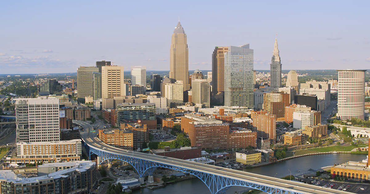 An aerial view of Cleveland, Ohio, featuring a bustling downtown skyline, iconic blue bridge, and clear skies. The urban landscape highlights the city's architecture and riverside setting on a sunny day.