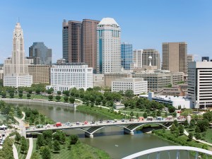 Panoramic view showcases downtown Columbus Georgia's modern architectural skyline, featuring glass skyscrapers alongside historic buildings, intersected by Scioto River and pedestrian bridges amid lush riverfront parks.