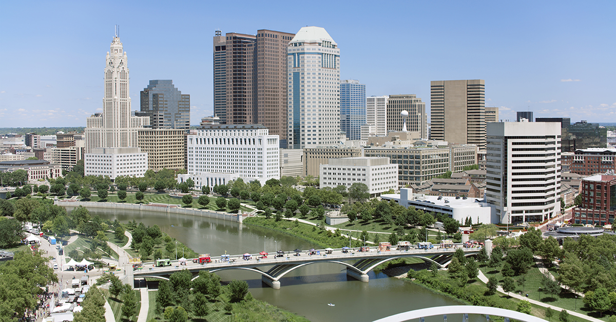 Panoramic view showcases downtown Columbus Georgia's modern architectural skyline, featuring glass skyscrapers alongside historic buildings, intersected by Scioto River and pedestrian bridges amid lush riverfront parks.