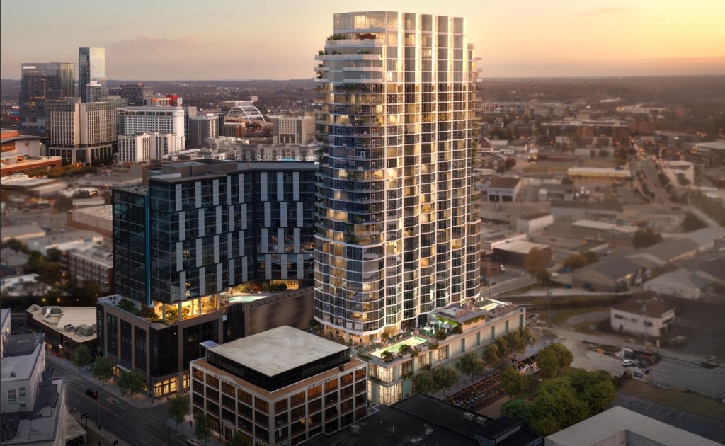 A modern cityscape at sunset featuring a tall, illuminated skyscraper with glass facades, surrounded by smaller urban buildings and lush greenery.