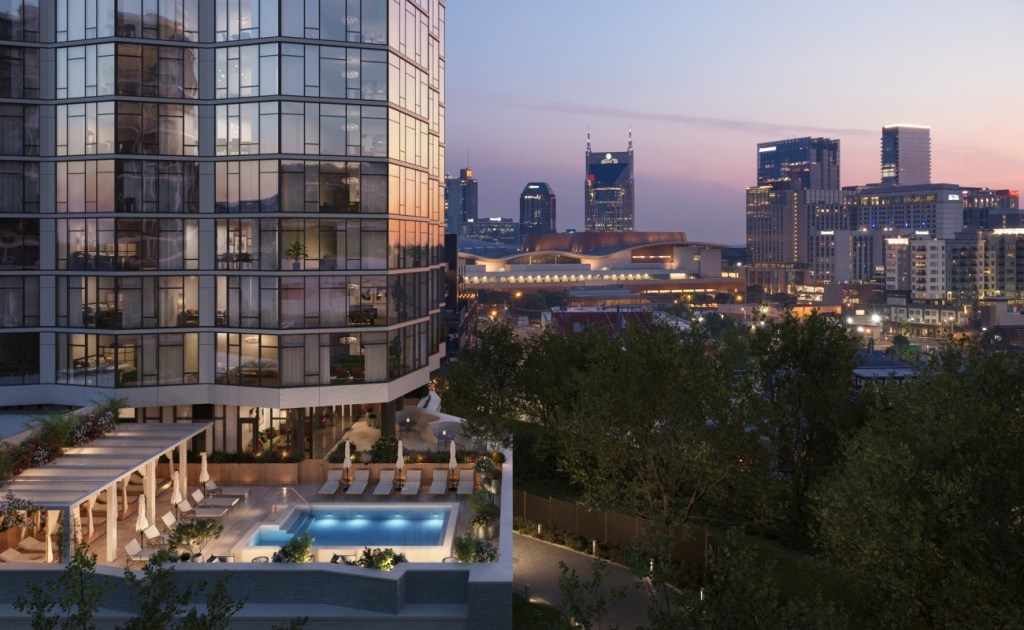 A modern cityscape at sunset featuring a tall, illuminated skyscraper with glass facades, surrounded by smaller urban buildings and lush greenery.