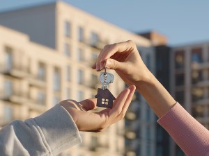 Close up of female real estate owner handing over house key with home shaped keychain to young man outdoors on background of modern multifamily apartment building.