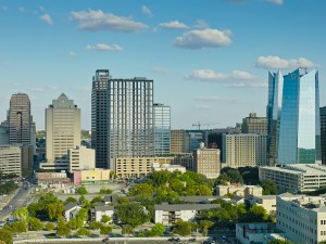 Drone shot of downtown San Antonio, Texas, featuring the city's modern skyline with a mix of high-rise office buildings, residential towers, and historic structures. The image captures the central business district with its distinctive blue glass towers and varied architectural styles, surrounded by tree-lined residential neighborhoods. The scene is photographed during daytime under partly cloudy skies, revealing the urban landscape that blends contemporary development with the city's established neighborhoods. Streets and parking areas are visible throughout the frame, illustrating the density and layout of this major Texas metropolitan area.