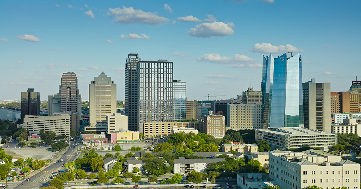Drone shot of downtown San Antonio, Texas, featuring the city's modern skyline with a mix of high-rise office buildings, residential towers, and historic structures. The image captures the central business district with its distinctive blue glass towers and varied architectural styles, surrounded by tree-lined residential neighborhoods. The scene is photographed during daytime under partly cloudy skies, revealing the urban landscape that blends contemporary development with the city's established neighborhoods. Streets and parking areas are visible throughout the frame, illustrating the density and layout of this major Texas metropolitan area.