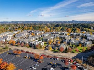 Aerial view of affordable housing community in Beaverton, Oregon. Several dozen three-story strucures with light gray roofs, against a background of trees turning fall colors and a low-lying mountain
