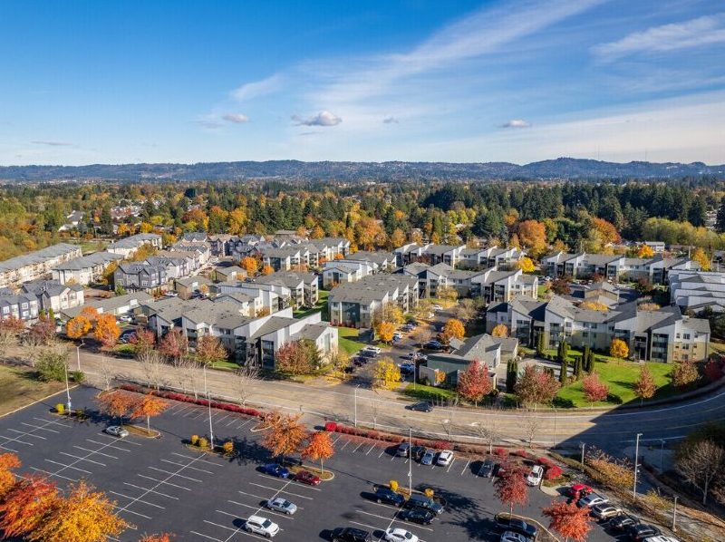Aerial view of affordable housing community in Beaverton, Oregon. Several dozen three-story strucures with light gray roofs, against a background of trees turning fall colors and a low-lying mountain