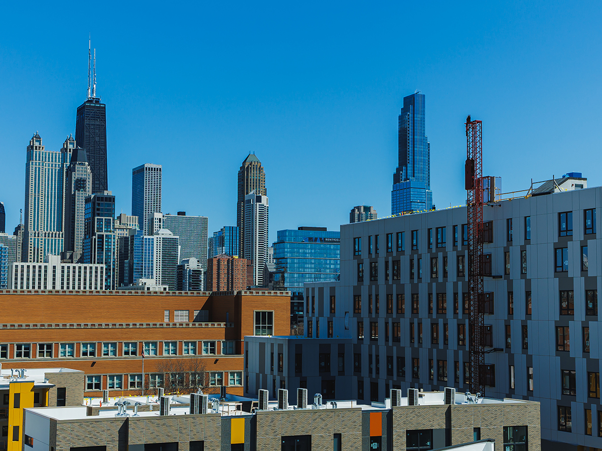 Tall buildings make up the skyline of Chicago. The scene shows a mix of architecture against a clear blue sky on a sunny day. The urban area has both new and old structures.
