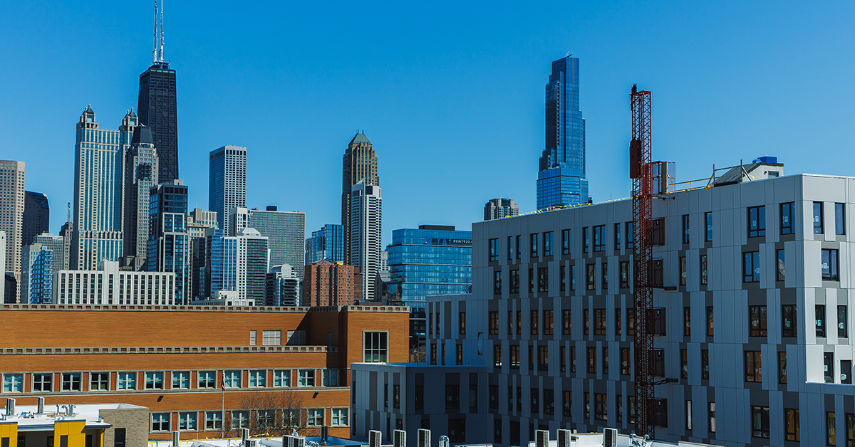 Tall buildings make up the skyline of Chicago. The scene shows a mix of architecture against a clear blue sky on a sunny day. The urban area has both new and old structures.