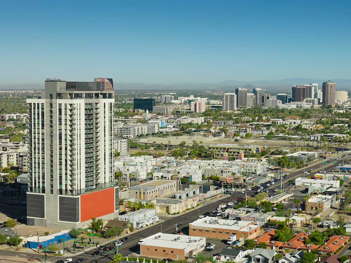 Aerial photograph of Midtown Phoenix, Arizona, with a modern high-rise residential tower in the foreground along Central Avenue. The Midtown business district rises in the middle distance with office towers, and the Phoenix Mountains are visible on the horizon under a clear blue sky. Low-rise residential neighborhoods, palm trees, and commercial buildings fill the urban landscape on a sunny day. • Authorization was obtained from the FAA for this operation in restricted airspace.