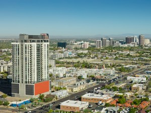 Aerial photograph of Midtown Phoenix, Arizona, with a modern high-rise residential tower in the foreground along Central Avenue. The Midtown business district rises in the middle distance with office towers, and the Phoenix Mountains are visible on the horizon under a clear blue sky. Low-rise residential neighborhoods, palm trees, and commercial buildings fill the urban landscape on a sunny day. • Authorization was obtained from the FAA for this operation in restricted airspace.