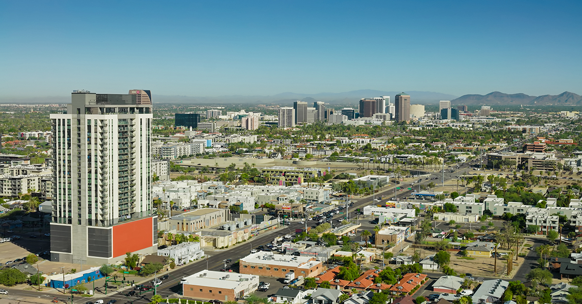 Aerial photograph of Midtown Phoenix, Arizona, with a modern high-rise residential tower in the foreground along Central Avenue. The Midtown business district rises in the middle distance with office towers, and the Phoenix Mountains are visible on the horizon under a clear blue sky. Low-rise residential neighborhoods, palm trees, and commercial buildings fill the urban landscape on a sunny day. • Authorization was obtained from the FAA for this operation in restricted airspace.