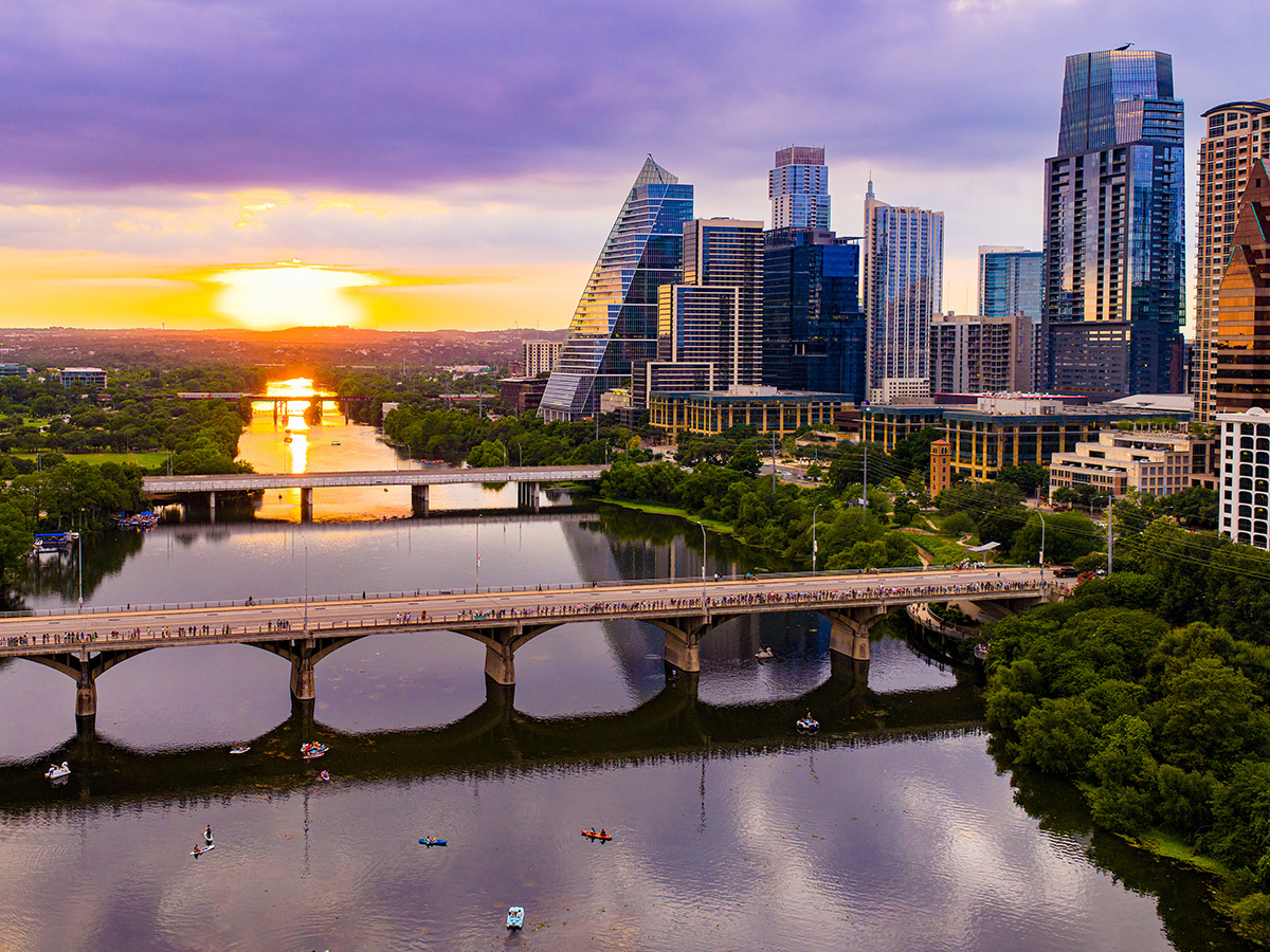 A stunning drone aerial view captures the Austin, Texas downtown skyline bathed in golden and purple sunset light, with the Colorado River (Lady Bird Lake) stretching beneath the Congress Avenue Bridge. Kayakers and paddleboarders dot the calm water below while crowds gather along the bridge to watch the spectacular summer sunset. The distinctive pyramid-shaped Frost Tower and surrounding modern glass skyscrapers reflect the warm evening light against a dramatic purple sky.