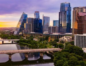 A stunning drone aerial view captures the Austin, Texas downtown skyline bathed in golden and purple sunset light, with the Colorado River (Lady Bird Lake) stretching beneath the Congress Avenue Bridge. Kayakers and paddleboarders dot the calm water below while crowds gather along the bridge to watch the spectacular summer sunset. The distinctive pyramid-shaped Frost Tower and surrounding modern glass skyscrapers reflect the warm evening light against a dramatic purple sky.